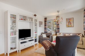 Ein Wohnzimmer mit Fernseher, weißen Regalen, einem grauen Sofa und einer gemusterten Decke.