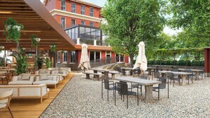 An outdoor seating area with tables and chairs, a building with a red facade, and a large tree providing shade.