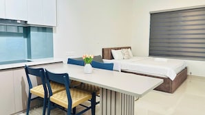 A modern hotel room with a dining table, two chairs, a bed with a headboard, a window with blinds, and a kitchenette. - SS Home Sunrise City Premium (Ho Chi Minh City)