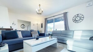 Un salon moderne avec un canapé bleu, un canapé blanc et une table basse. On y trouve un lustre, une horloge et une fenêtre avec des rideaux.