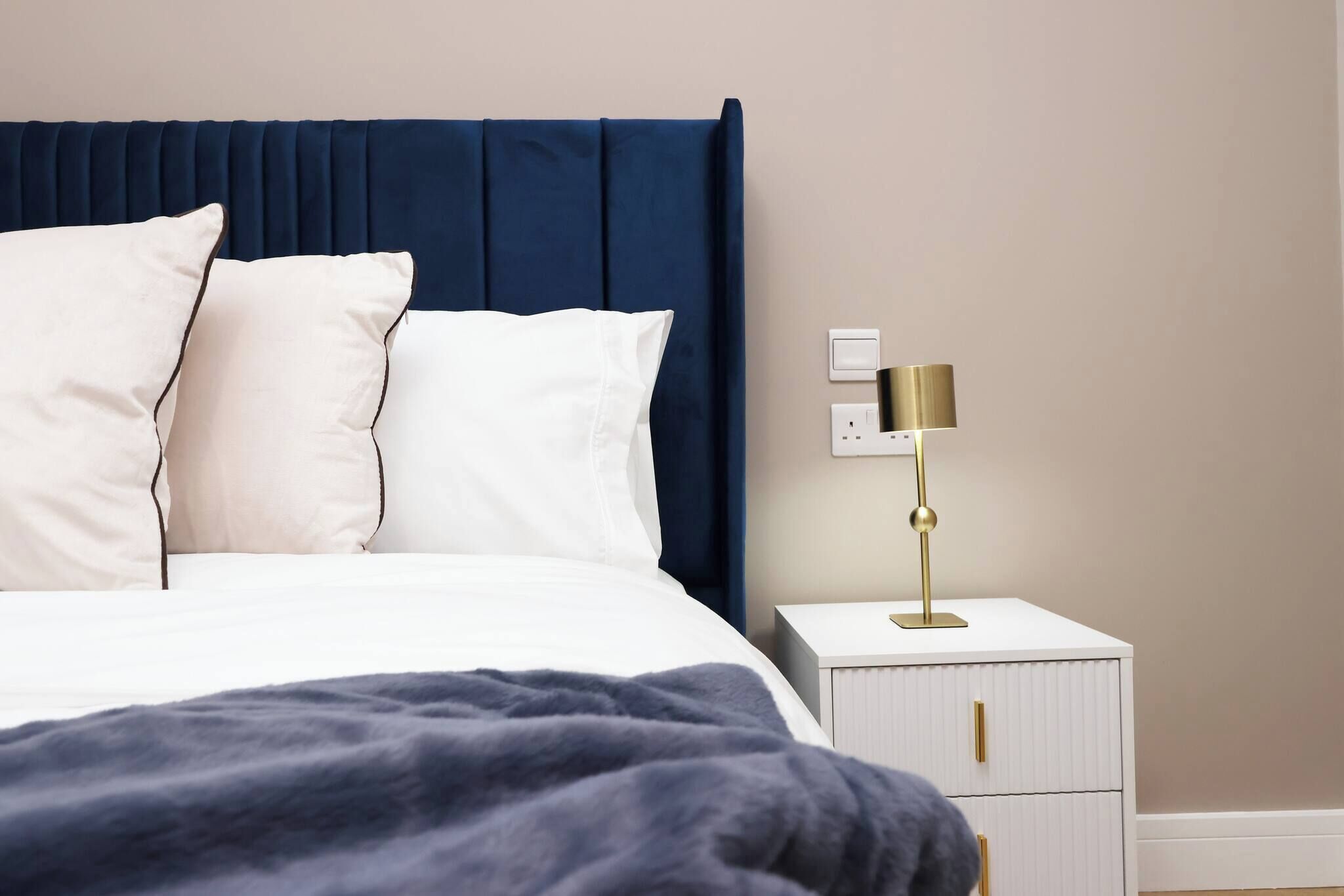 A bed with a dark headboard, white bedding, and a blue blanket. There is a bedside table with a lamp and a power outlet.