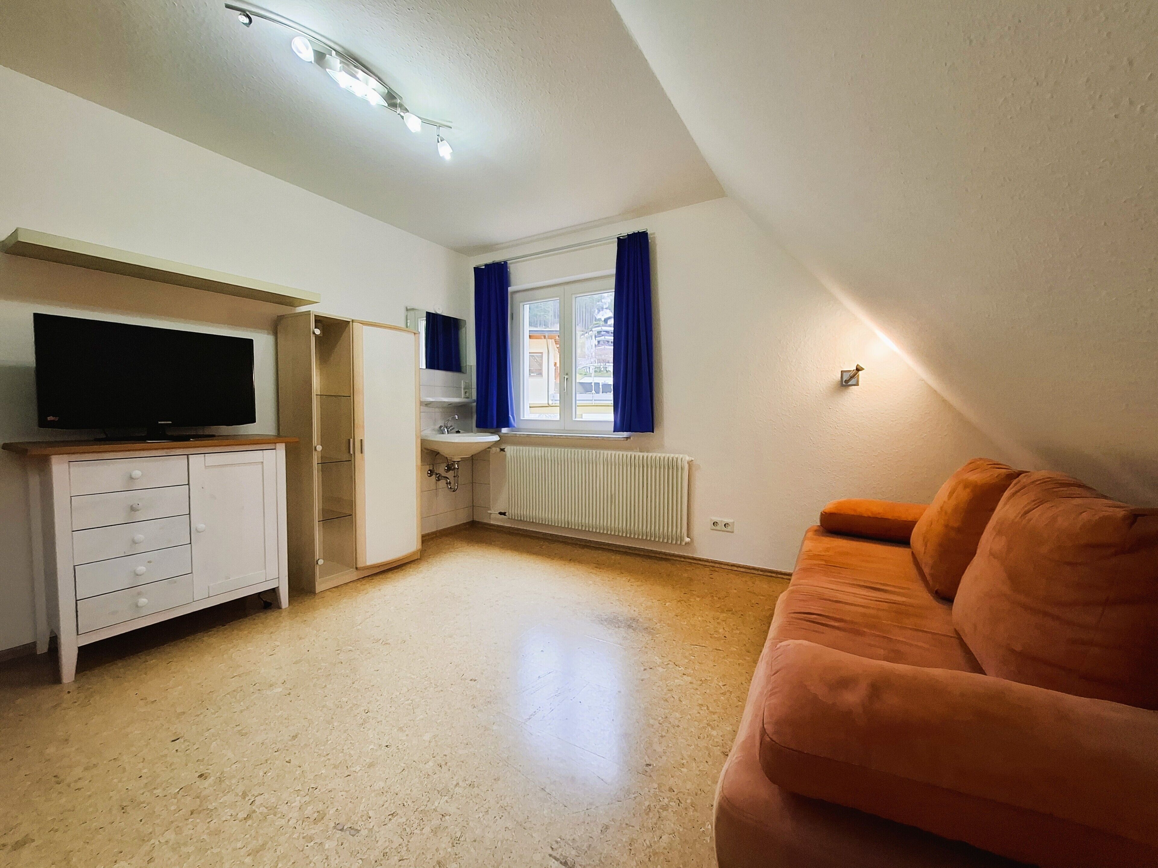 A room with a television, a white cabinet, a sink, a window with blue curtains, and an orange couch.