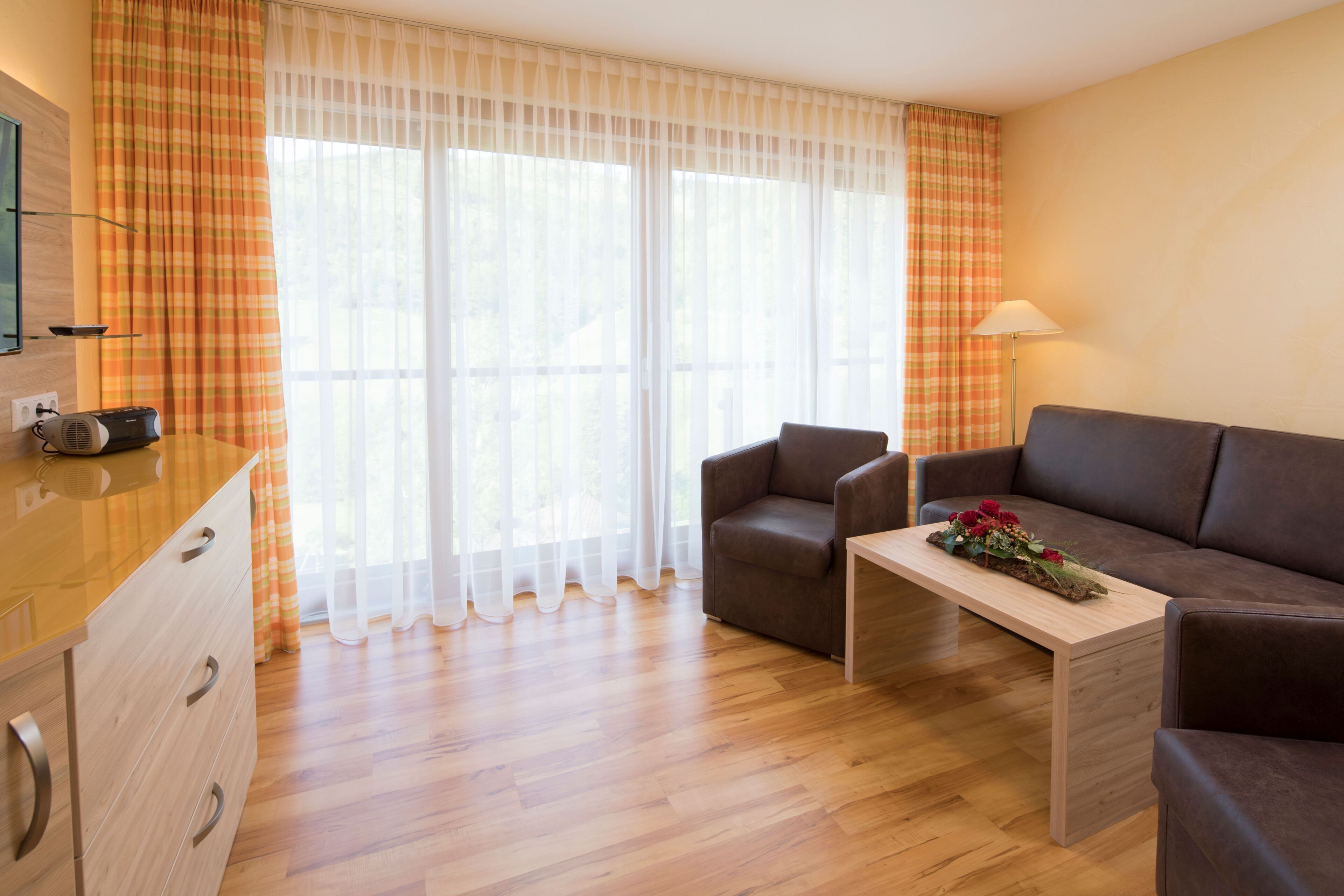 Un salon moderne avec un plancher en bois, un canapé marron, une table basse en bois agrémentée d’un arrangement floral, un téléviseur posé sur un meuble en bois et des rideaux légers.