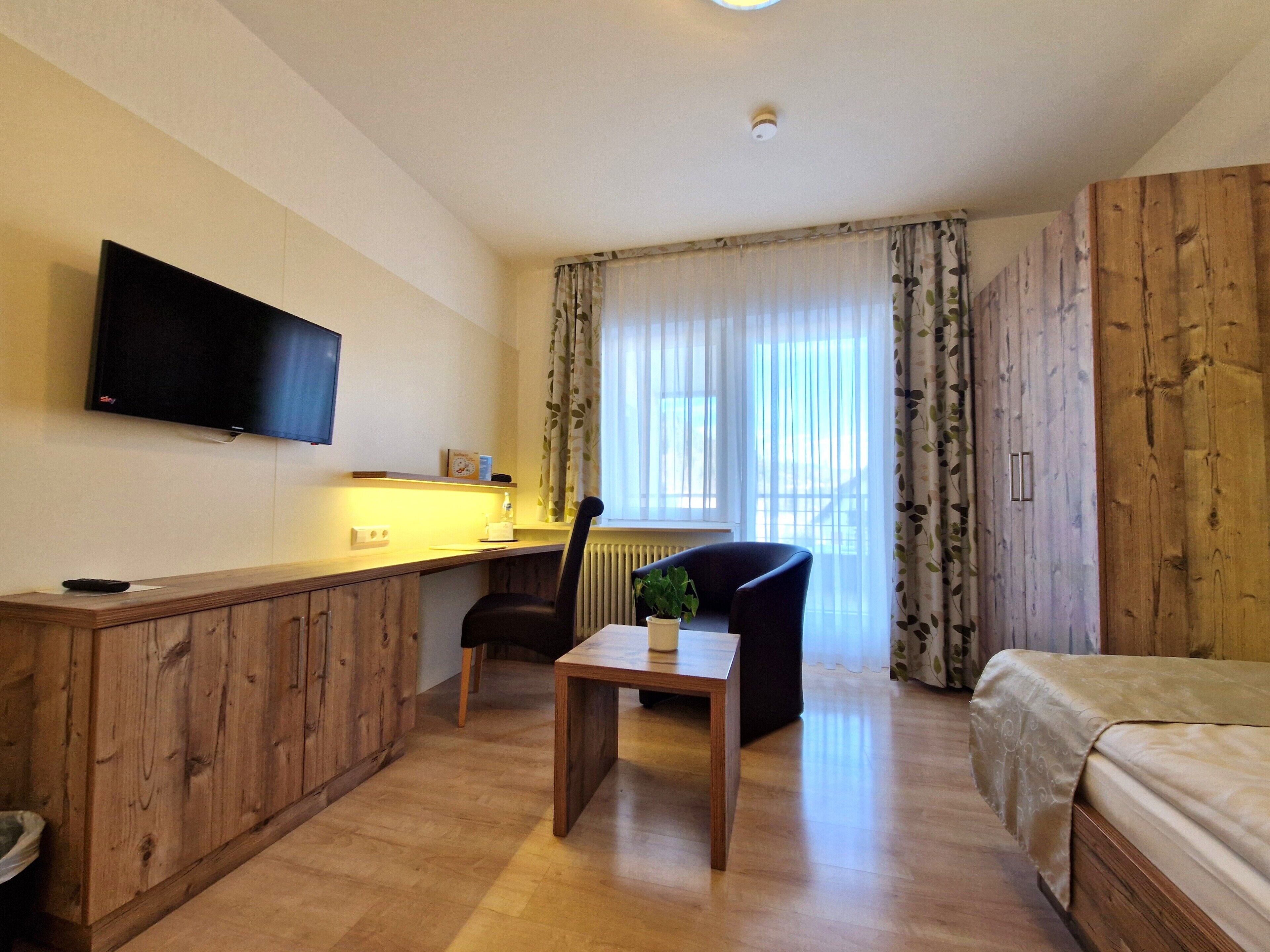 Een hotelkamer met een houten bureau, een stoel, een bed, een aan de muur bevestigde televisie en een raam met gordijnen.