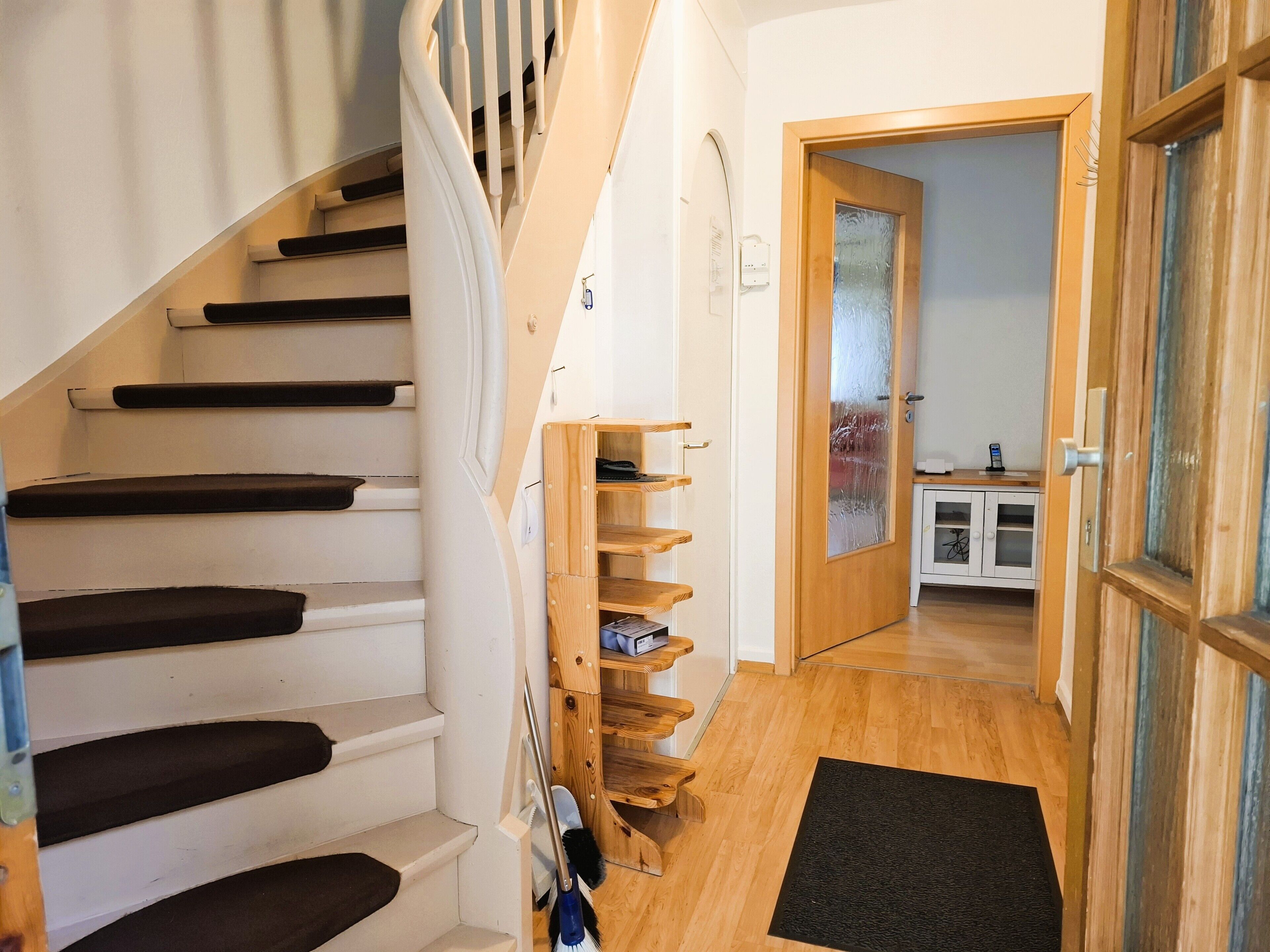 Un escalier avec des marches noires, un banc en bois doté d’un espace de rangement, une porte en verre dépoli et, au fond, un meuble blanc.