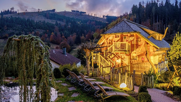 A wooden cabin with a deck, surrounded by a pond, trees, and a hilly landscape. - Tanne Tonbach - Wellness & Wohlfühl Hotel (Baiersbronn)