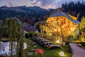 A wooden cabin with a deck, surrounded by a pond, trees, and a hilly landscape. - Tanne Tonbach - Wellness & Wohlfühl Hotel (Baiersbronn)