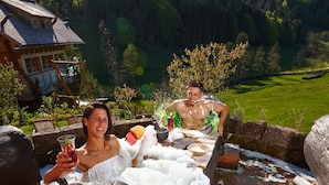A wooden outdoor bathtub with a rustic setting, surrounded by stone walls and a forested hillside. - Tanne Tonbach - Wellness & Wohlfühl Hotel (Baiersbronn)