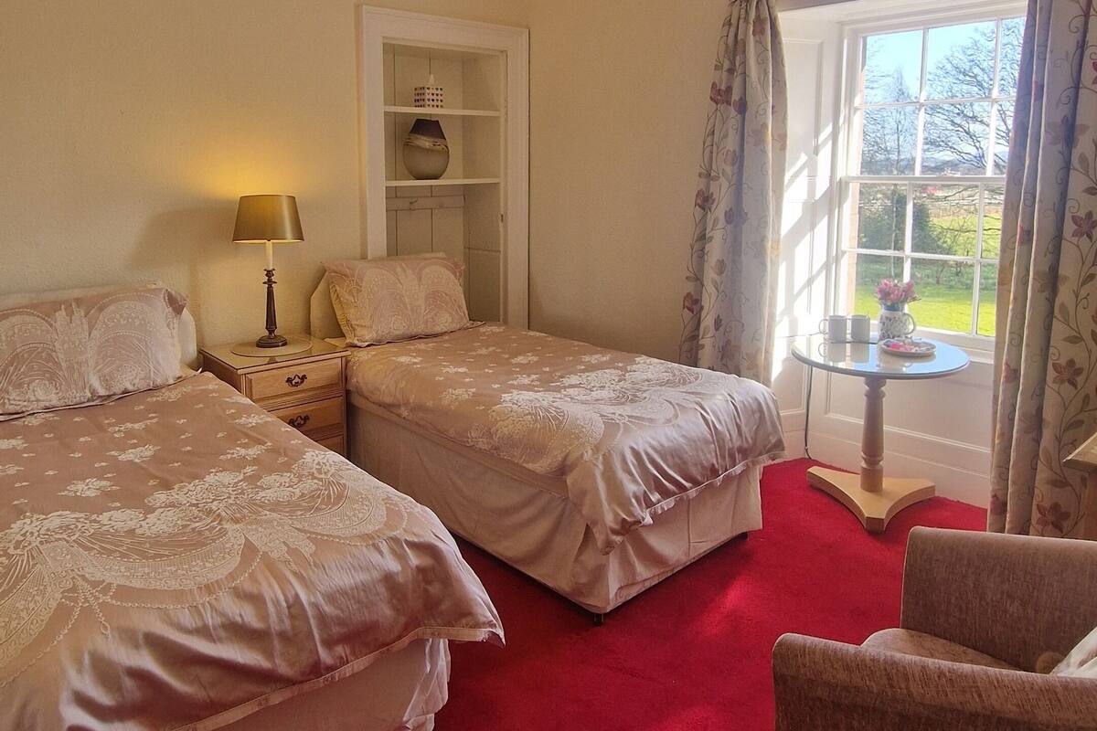 A hotel room with two beds, a red carpet, a chair, a table with a vase, and a window with curtains.