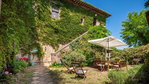 A stone building with ivy-covered walls, a stone pathway, wooden furniture, and a table with an umbrella. - Apartment at Tenuta Bichi Borghesi Farm (Monteriggioni)
