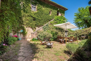 A stone building with ivy-covered walls, a stone pathway, wooden furniture, and a table with an umbrella. - Apartment at Tenuta Bichi Borghesi Farm (Monteriggioni)