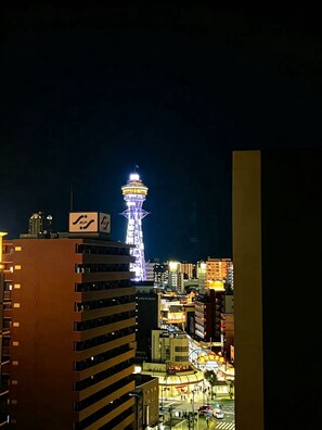 Un paisaje urbano nocturno con una torre destacada iluminada en azul y amarillo, rodeada de varios edificios y una calle concurrida.