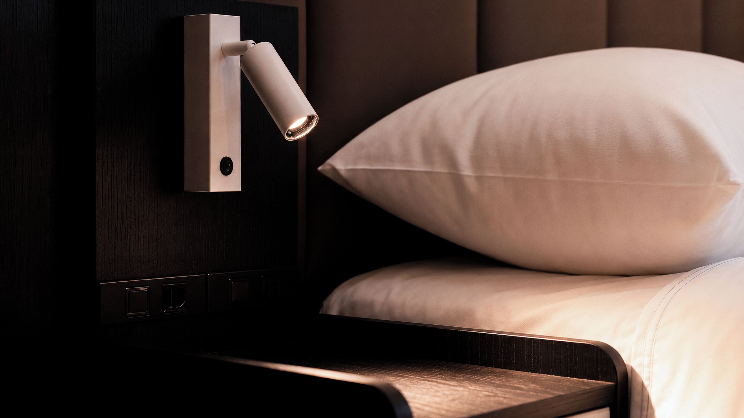 A bed with a white pillow and a bedside table with a lamp.