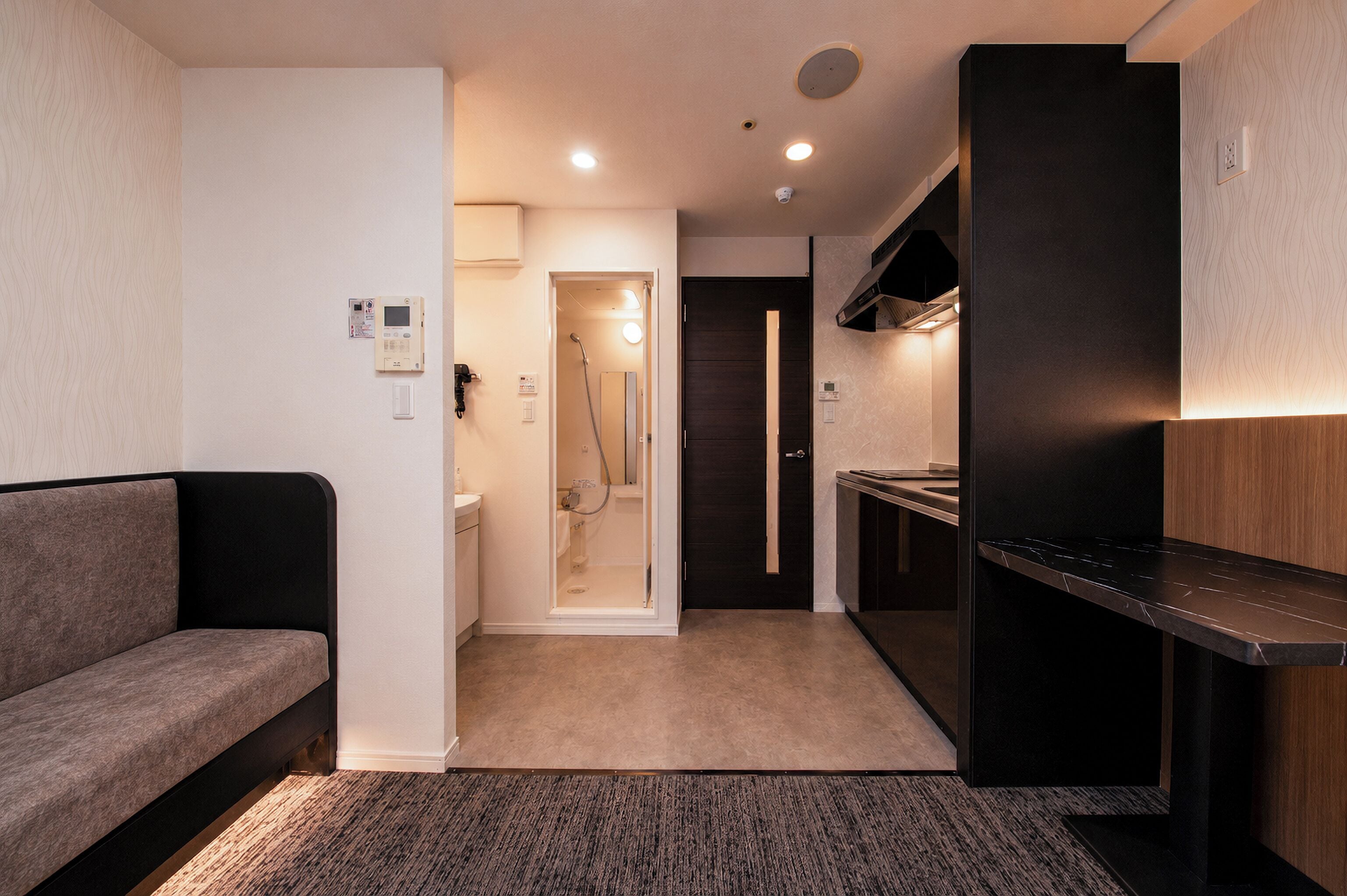 Een compacte hotelkamer met een bank, een kitchenette, een douchegedeelte en een deur naar een andere kamer.