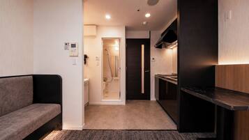 Een compacte hotelkamer met een bank, een kitchenette, een douchegedeelte en een deur naar een andere kamer.