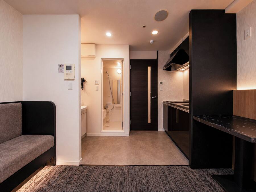 Een compacte hotelkamer met een bank, een kitchenette, een douchegedeelte en een deur naar een andere kamer.