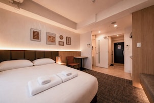 Quarto de hotel com uma cama grande, uma mesa de cabeceira, uma cadeira, uma mesinha e uma porta para o banheiro.