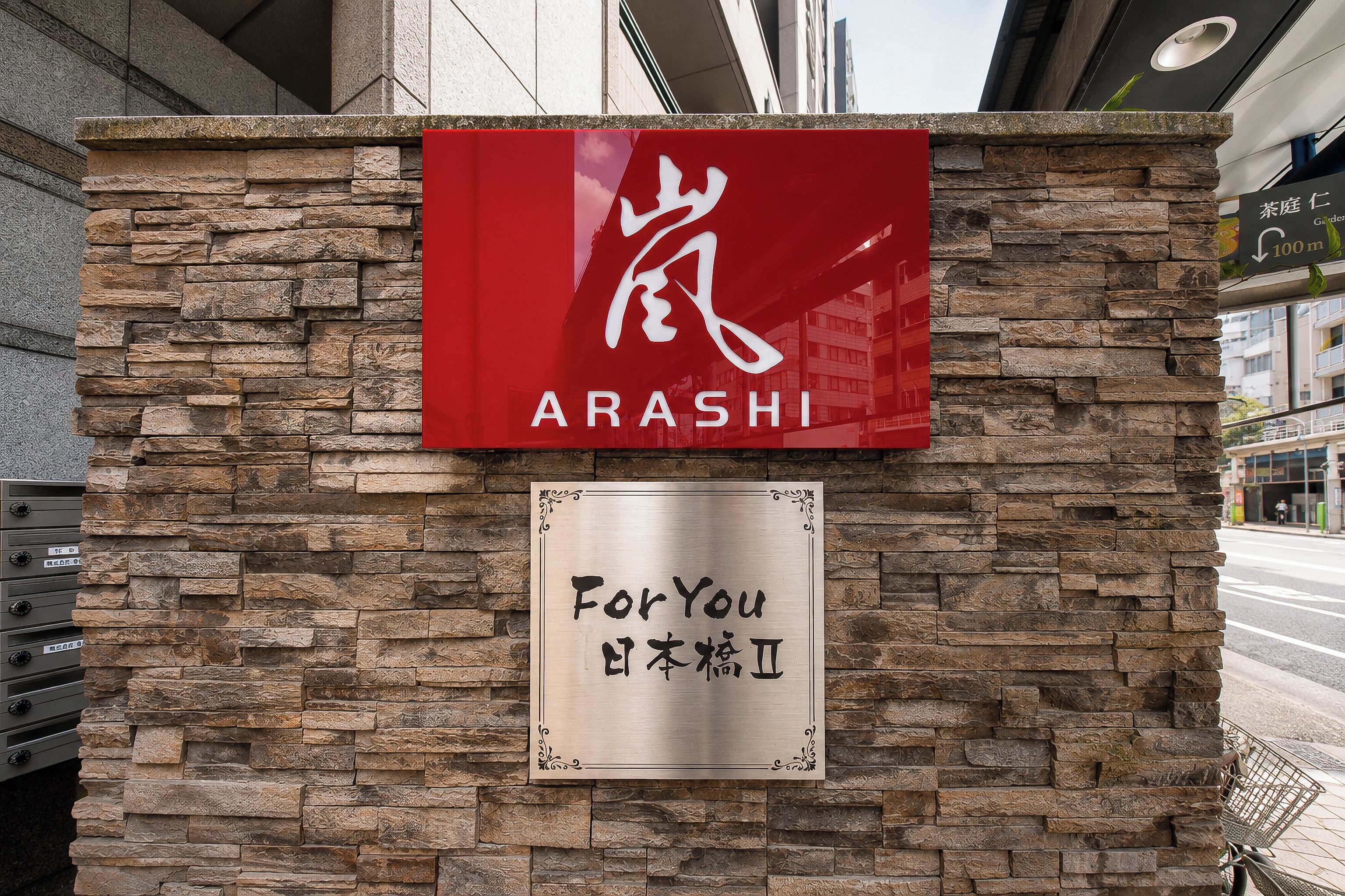 Uma parede de pedra com uma placa vermelha com a inscrição 'ARASHI' e uma placa com a frase 'For You' e caracteres chineses.