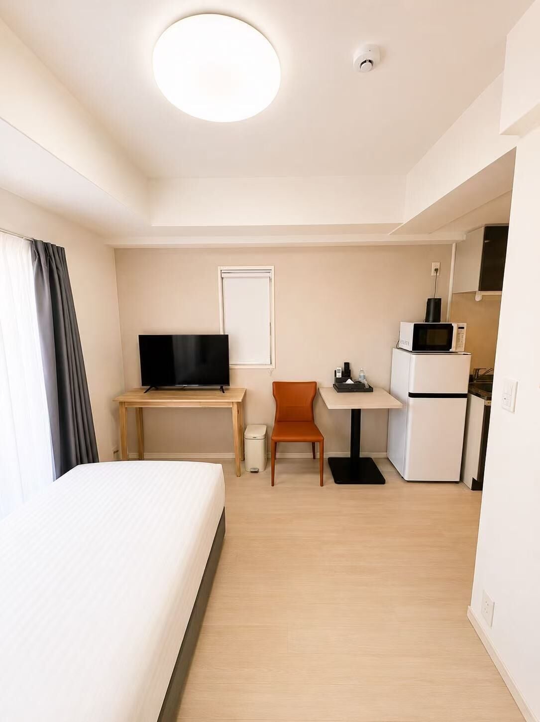 A hotel room with a bed, a desk with a flat-screen TV, a chair, a small kitchenette with a microwave and refrigerator, and a ceiling light.