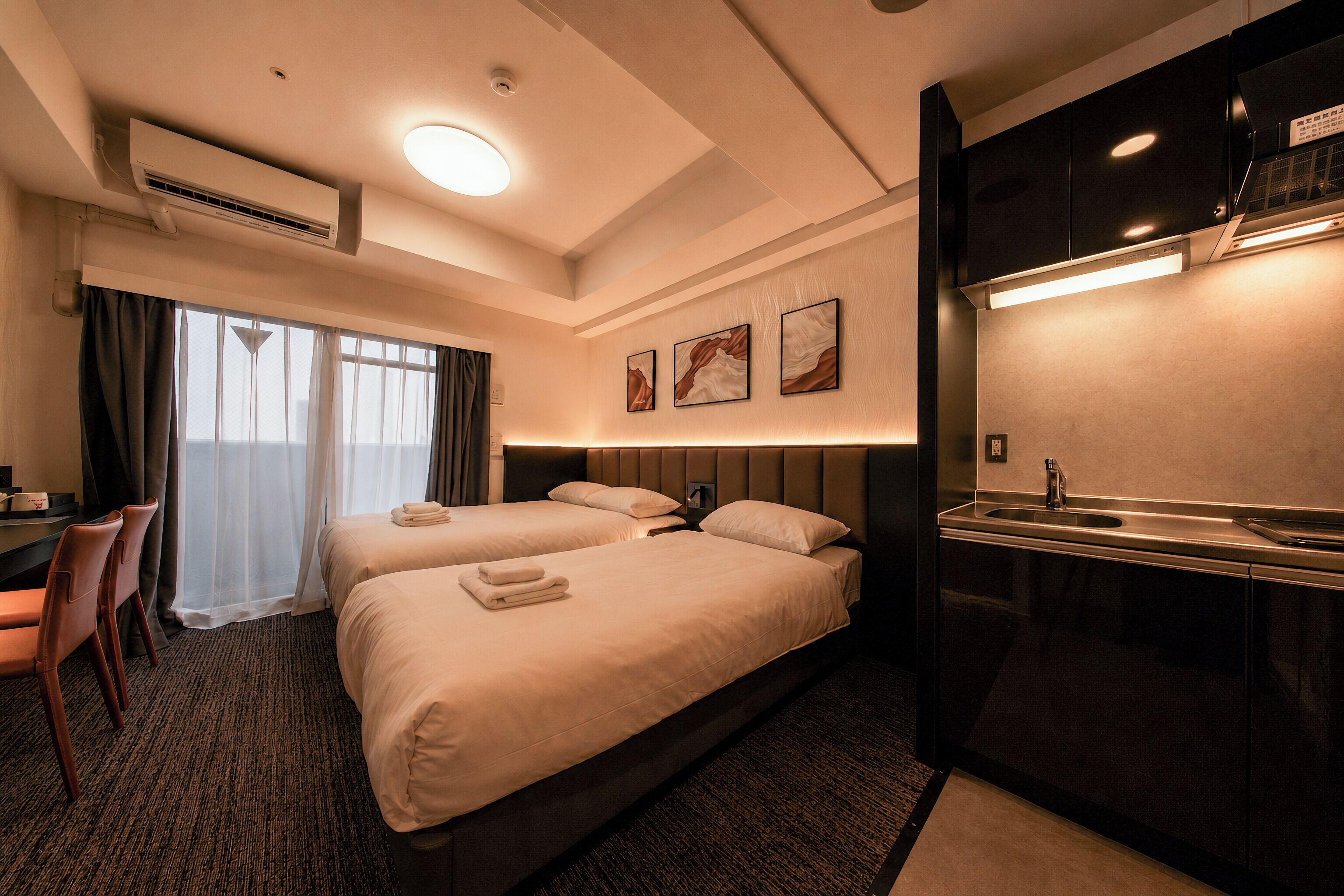 Habitación de hotel con dos camas, un escritorio, una silla, un baño con lavamanos y obras de arte en la pared.