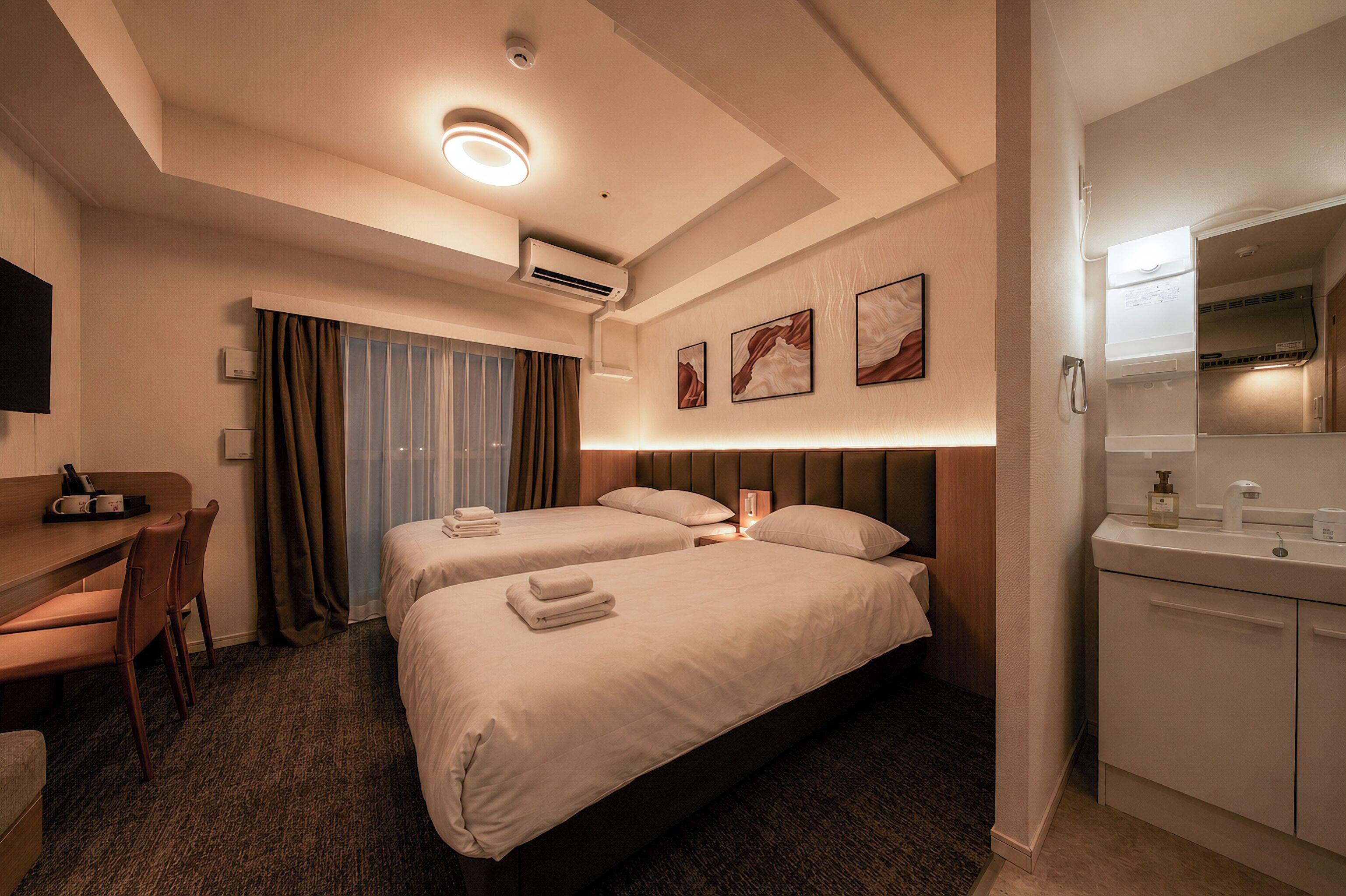 A hotel room with two beds, a desk, a chair, a bathroom, and a flat-screen TV.