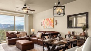 A living room with a brown leather sofa, a dining table with chairs, a ceiling fan, a chandelier, a large window with a mountain view, and artwork on the wall. - Sedona Getaway Sedona! 2-King, Hiking, Views, Pets! (Sedona)