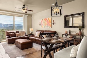 A living room with a brown leather sofa, a dining table with chairs, a ceiling fan, a chandelier, a large window with a mountain view, and artwork on the wall. - Relaxing Sedona Hiking HQ, View, West Sedona! Star (Sedona)