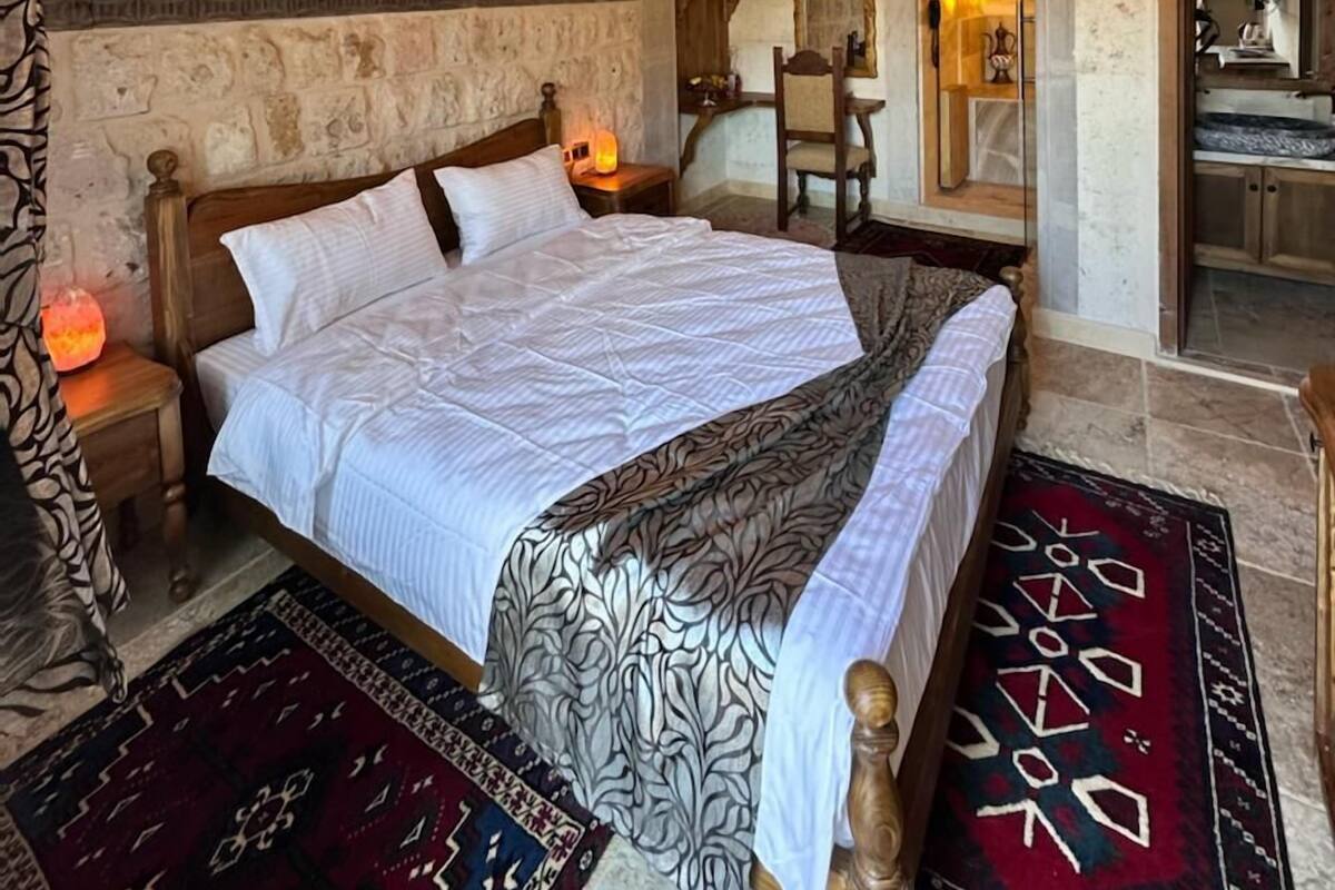 Un dormitorio con techo de madera, un candelabro, una cama con ropa blanca, una pared de piedra y una mesita con lámpara.