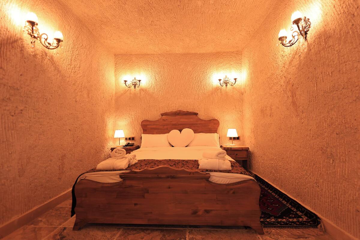 Una habitación con una cama de madera, almohadillas con forma de corazón, dos lámparas fijadas a la pared y una mesita de noche con lámpara.