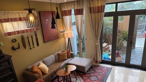A living room with a sofa, a coffee table, and a patterned rug. There is a large window with curtains and a view of the outside. - An Bang Antique Beach House (Đà Nẵng)