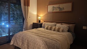 A bedroom with a large bed, a nightstand, a lamp, a window with curtains, and a framed artwork on the wall. - An Bang Antique Beach House (Đà Nẵng)