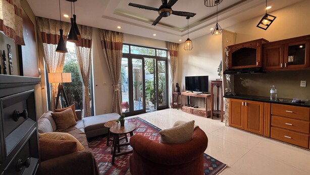 A spacious living area with a sofa, a coffee table, and a television. There is a kitchen area with wooden cabinets and a countertop. - An Bang Antique Beach House (Đà Nẵng)