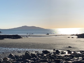A beach with rocks in the foreground, a calm sea, and a mountain in the distance.
