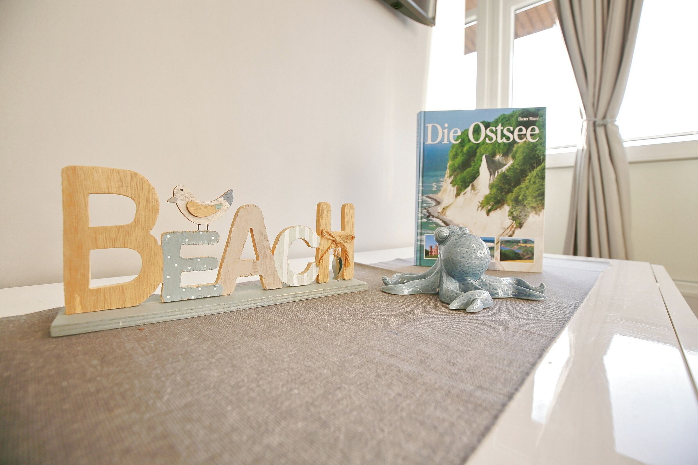 Una habitación con un letrero de madera que dice 'BEACH', un adorno de pájaro y un libro 'Die Ostsee' sobre una mesa.
