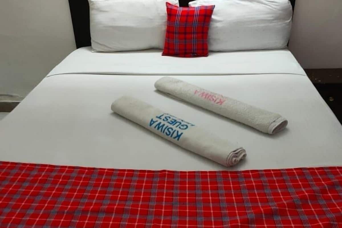 A hotel room with a bed, white bedding, a red plaid blanket, and a rolled towel with text.