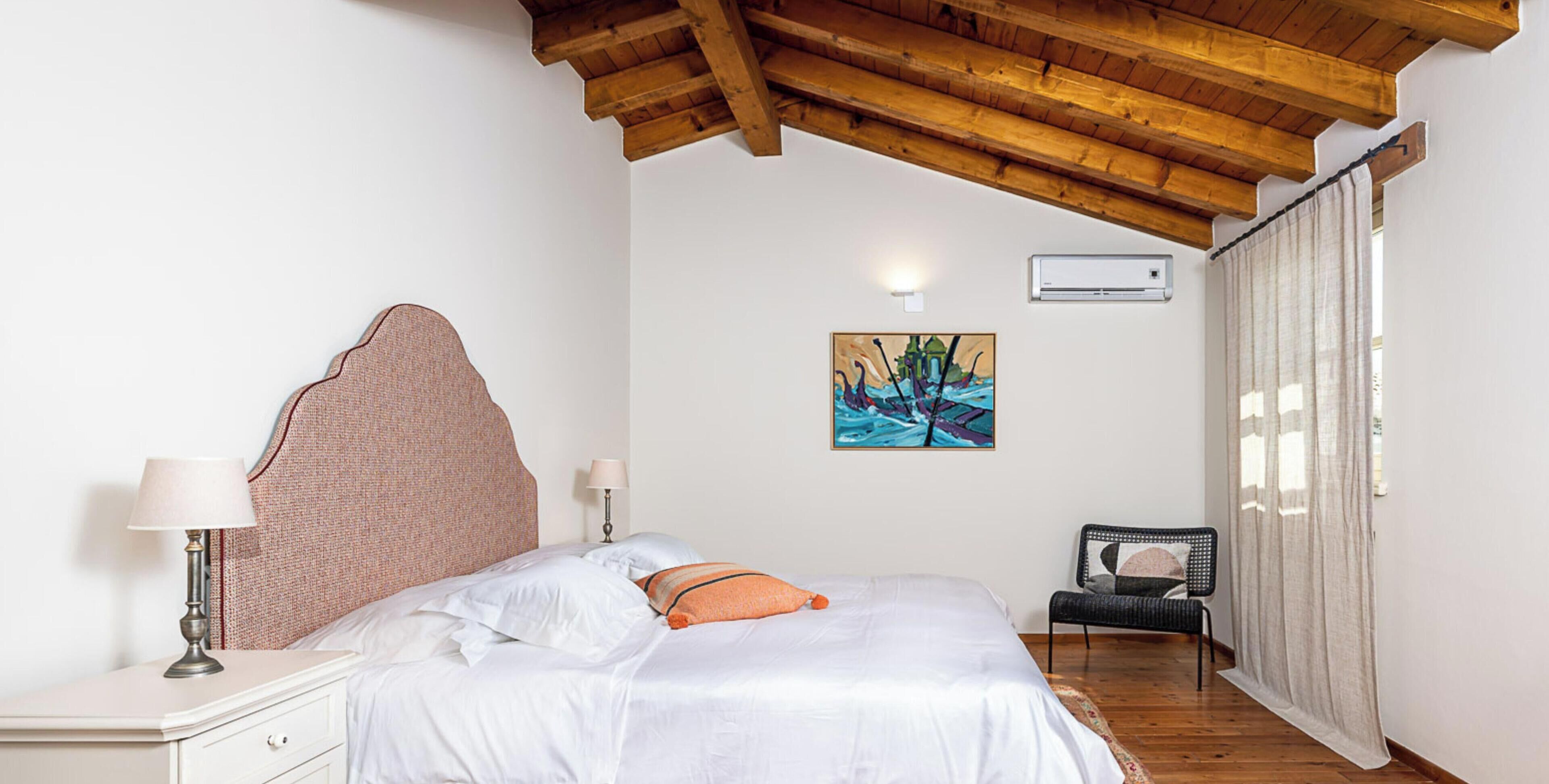 A bedroom with a wooden ceiling, a bed with white bedding, a nightstand with a lamp, a chair, and a painting on the wall.