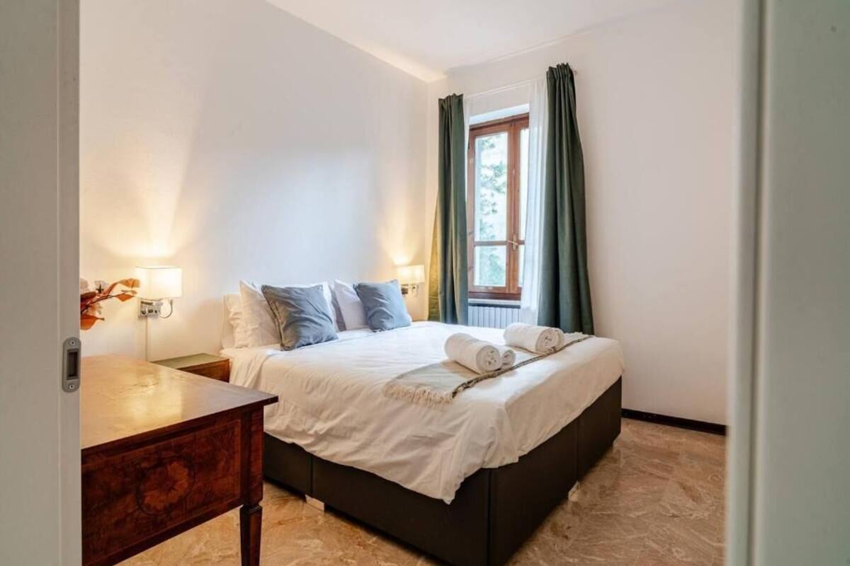 A neatly made bed with white linens and a dark headboard, a bedside table with a lamp, and a window with green curtains.