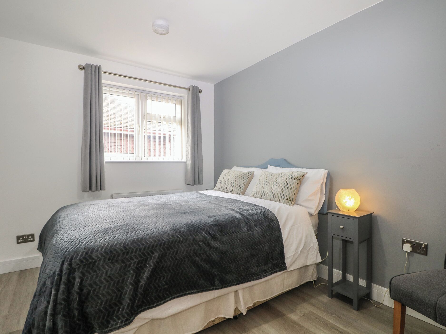 Une chambre à coucher avec un lit, une table de chevet avec une lampe, une fenêtre avec des rideaux et une prise électrique au mur.
