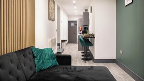 Una sala de estar moderna con un sofá negro y un cojín verde, una zona de cocina con taburetes de bar y una mesa de comedor.