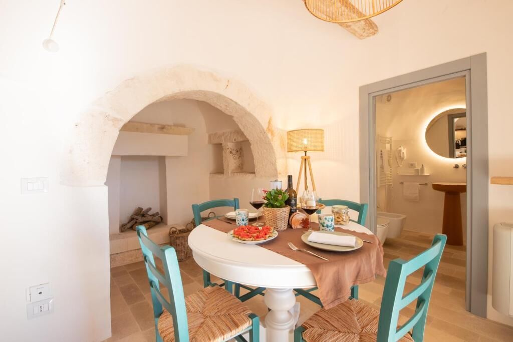 A dining area with a round table, teal chairs, and a white tablecloth. There is a lamp, a bottle of wine, and a basket with food. An archway leads to another room with a mirror and a sink.
