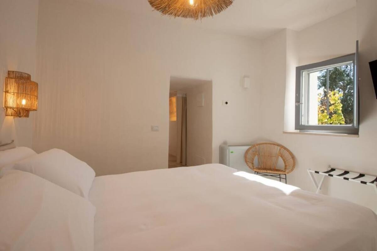 A bedroom with a bed, a wicker chair, a TV mounted on the wall, and a window with a view of trees.