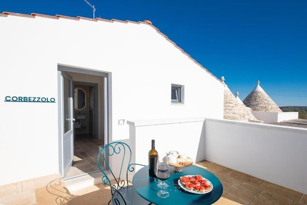 A white building with a sign reading "CORBEZZOLO" and a balcony with a table set for two, a bottle of wine, and a plate of food.