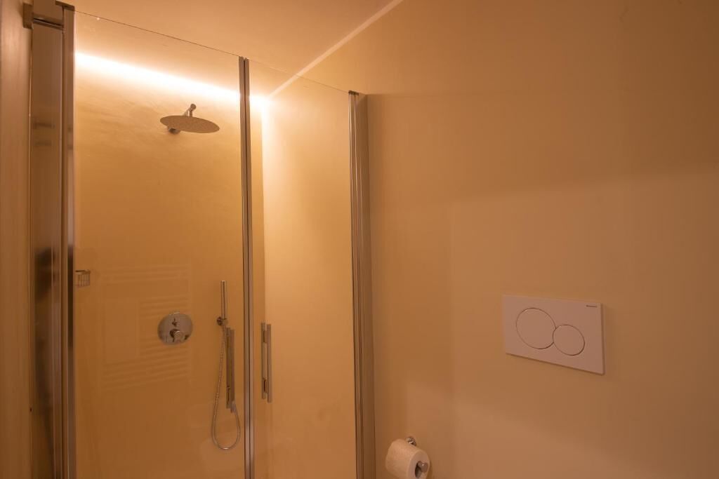 Una ducha con mampara de vidrio, cabezal de ducha de mano y dispensador de papel higiénico en la pared.