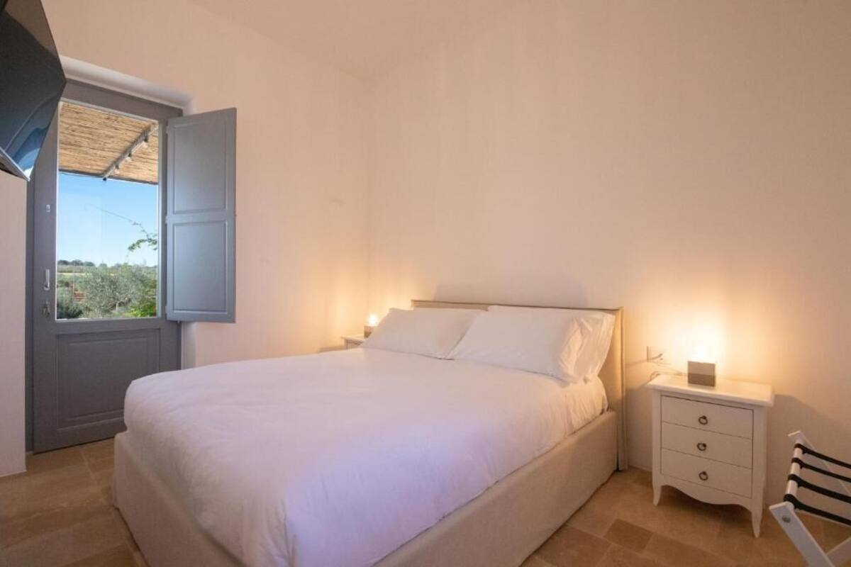 A bedroom with a bed, a nightstand, a chair, and a window with a view of greenery.