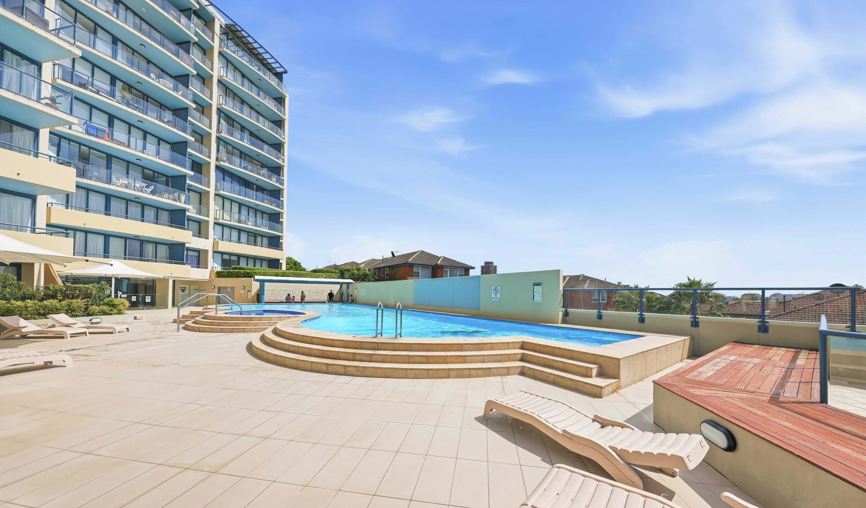 En pool på taket med ett avslappningsområde vid poolkanten, en byggnad med balkonger och en klar himmel.