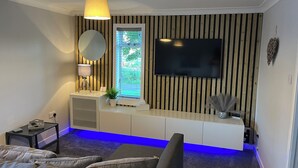 Ein modernes Wohnzimmer mit flachbildfernseher an der Wand, einem runden Spiegel, einem kleinen Tisch mit Lampe und einem Sofa.