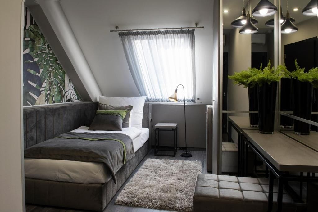 A compact bedroom with a bed, a small desk, a mirror, and a window with sheer curtains.