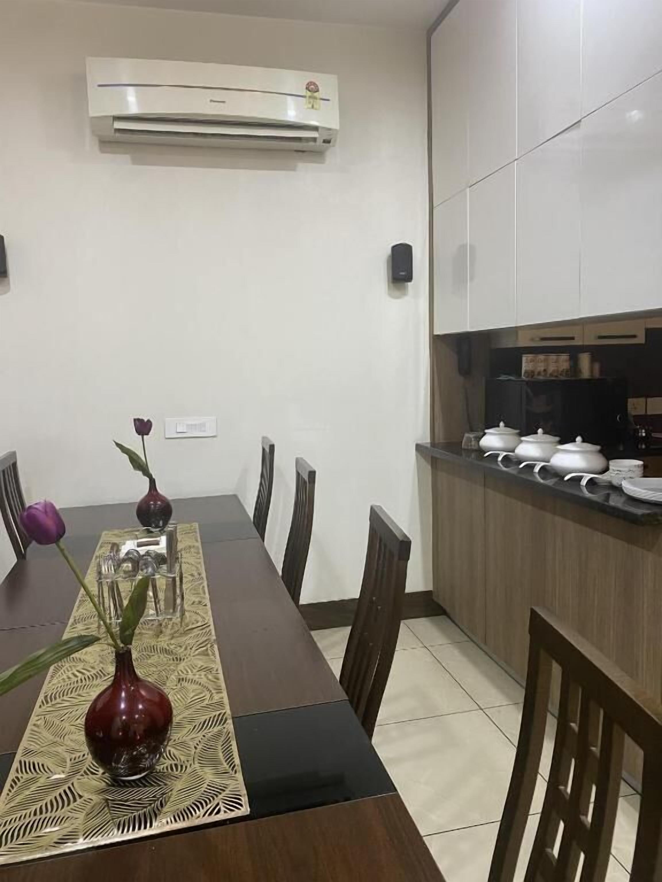 Un comedor con una mesa de madera larga, sillas y un florero con flores. Hay una zona de cocina con armarios y un refrigerador.