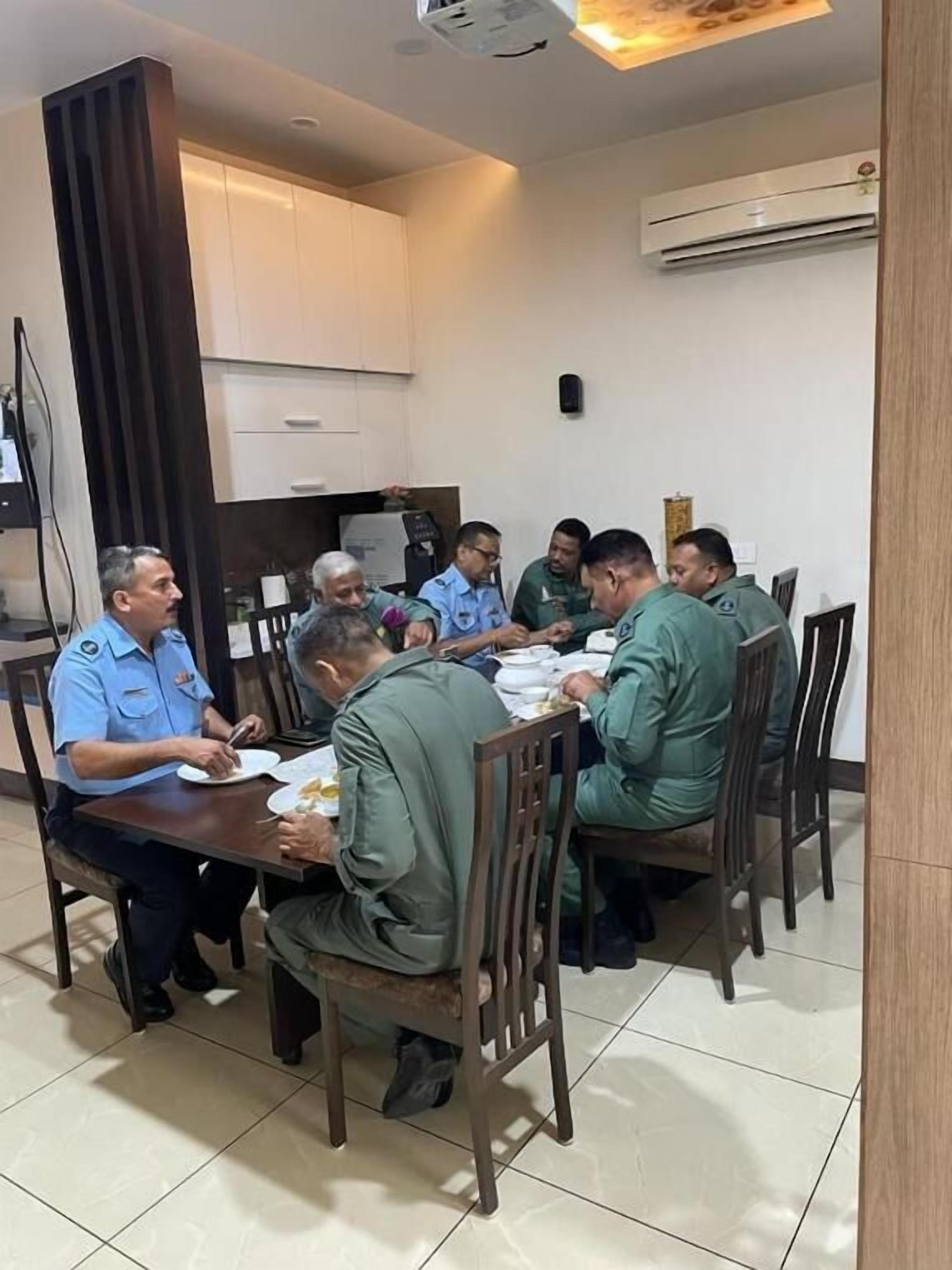 Um grupo de pessoas sentadas à volta de uma mesa numa sala com soalho em azulejo, uma luminária no teto e um aparelho de ar condicionado na parede.