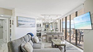 A modern living room with a grey sofa, a dining area with chairs, a kitchen with a large island, and a balcony with a TV. - Gulf-Front Luxury | Surfside 11th Floor | #1109 | 2B/2Ba (Miramar Beach)